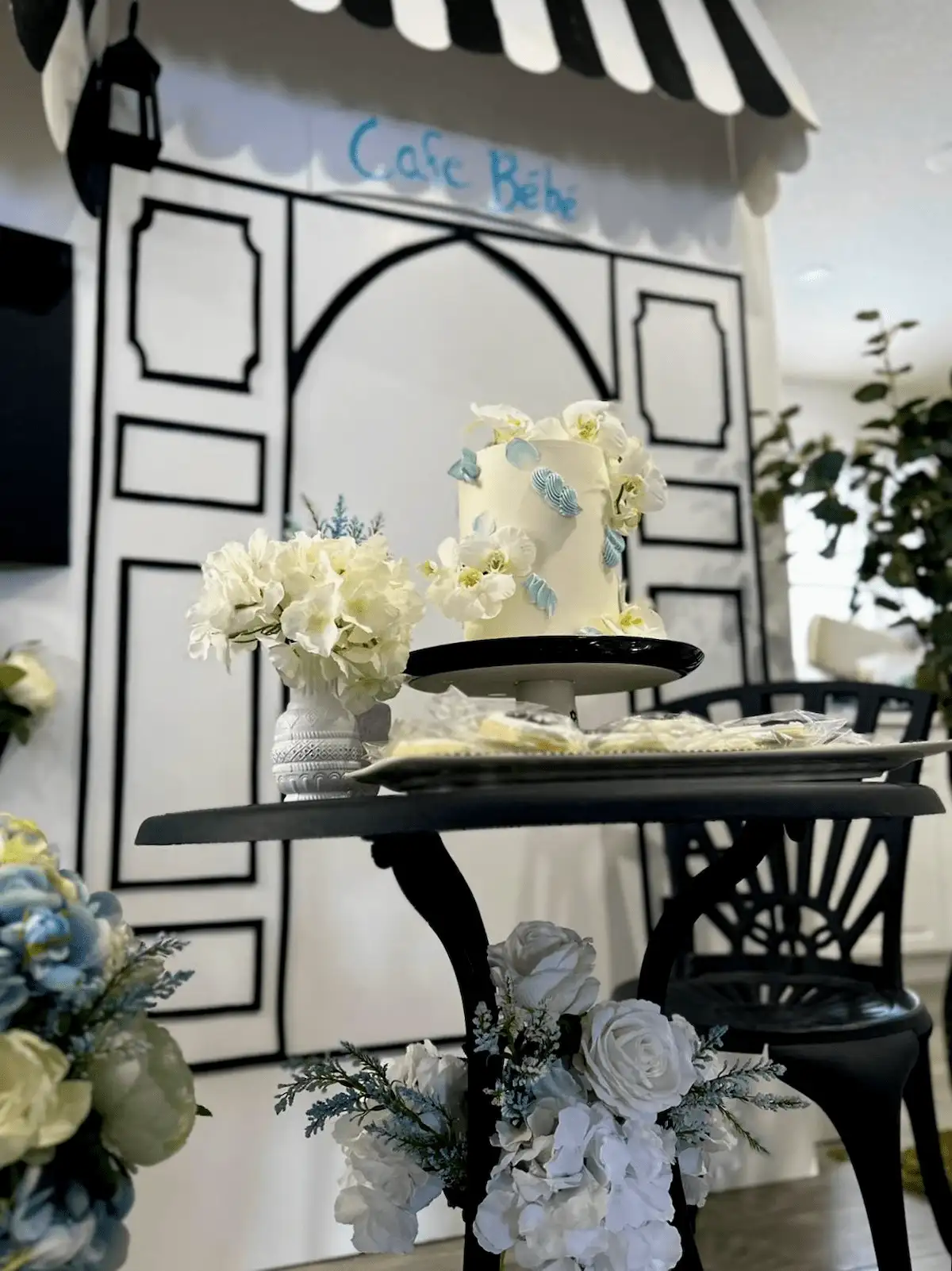 A close-up of a Parisian café-inspired dessert table featuring a white and blue floral-themed cake with delicate sugar flowers, elegant cookies, and a vase of white hydrangeas, set against a black bistro table adorned with floral arrangements.