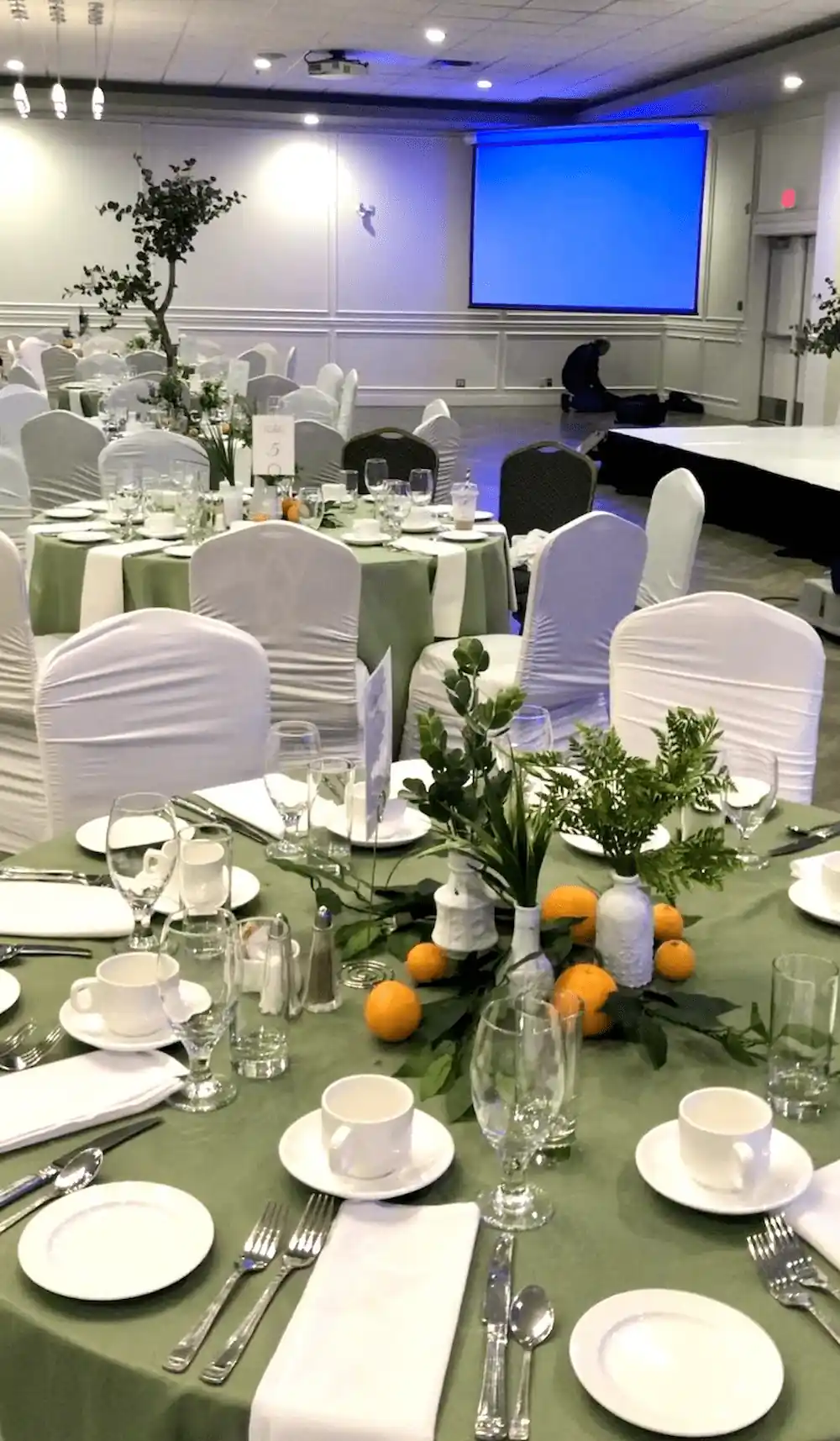  A banquet hall set for an elegant event, featuring round tables draped in sage green tablecloths with white dinnerware, silverware, and glassware. The centerpiece consists of fresh greenery, small white vases, and scattered oranges, creating a natural and sophisticated ambiance.