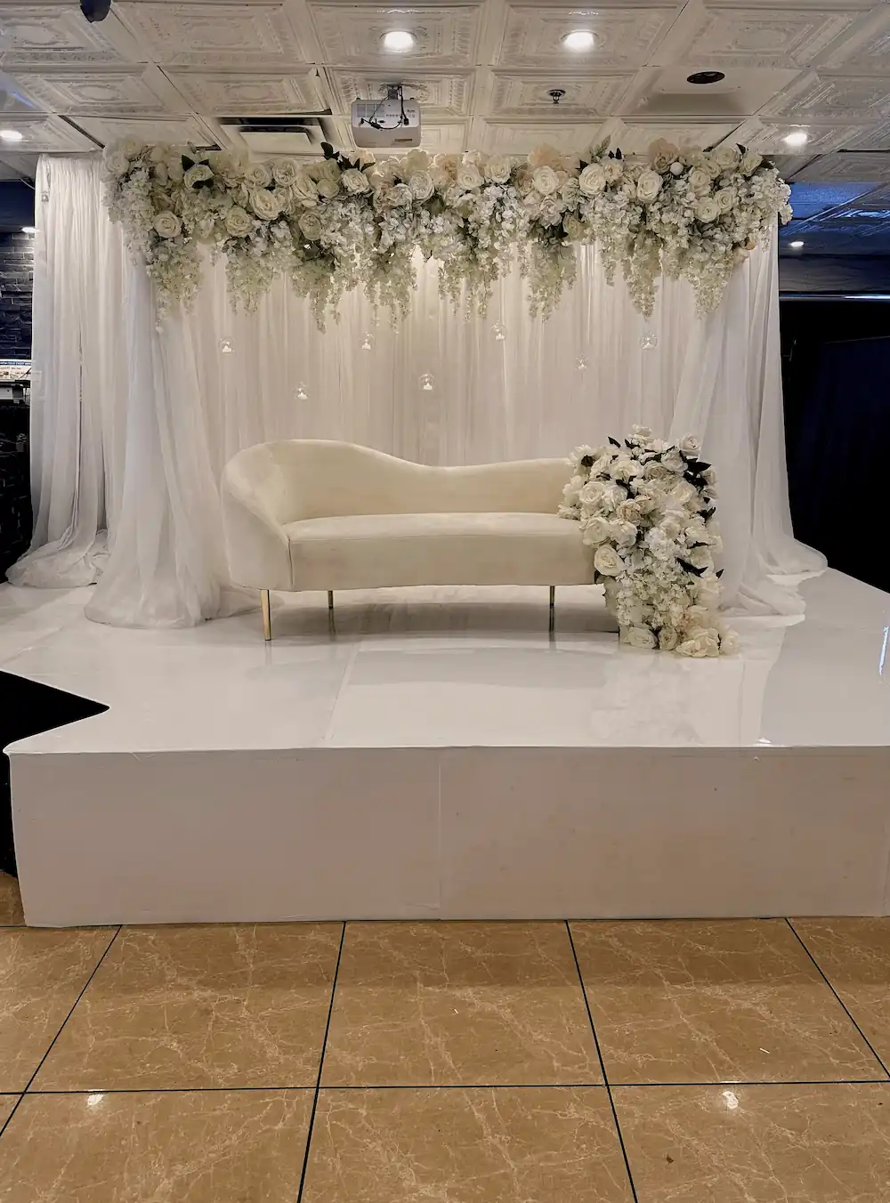 A grand event stage with an ivory chaise lounge set on a raised platform, draped with flowing white curtains. The backdrop features cascading white roses and hydrangeas with hanging glass candle orbs, creating a romantic and luxurious setting.