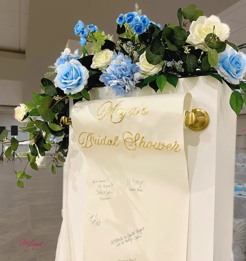 Close-up of a bridal shower guestbook scroll with gold calligraphy, surrounded by a floral arrangement of soft blue and white roses. A creative and elegant way for guests to leave messages at a wedding or bridal event.