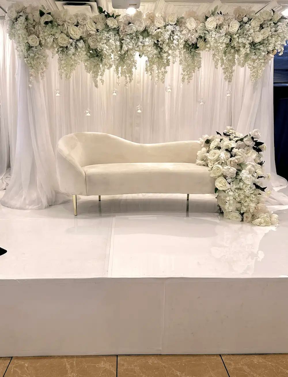 A close-up of the event stage, highlighting the elegant white floral arrangements, the plush ivory chaise lounge, and the delicate glass candle orbs suspended in front of the soft white drapery.