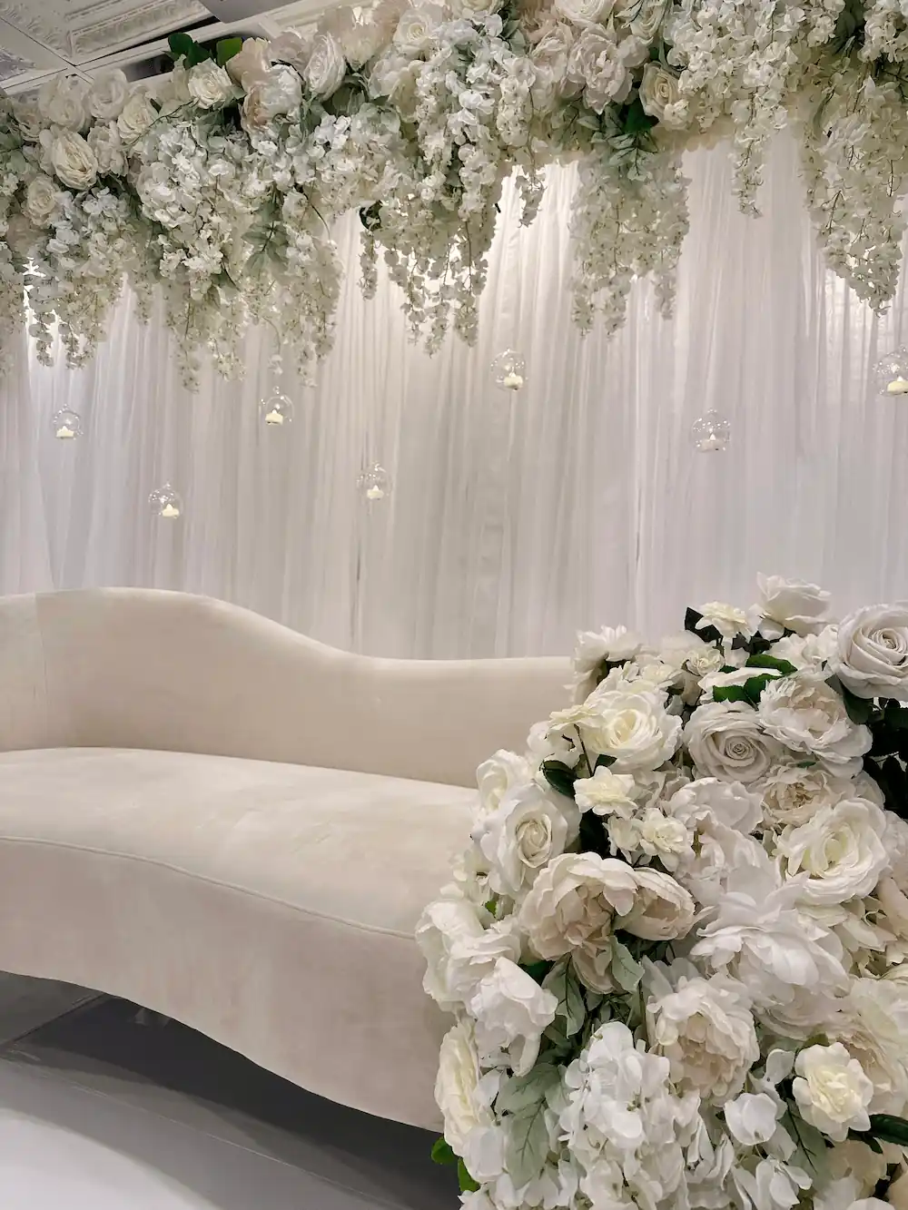 A detailed close-up of the floral arrangements on the event stage, featuring cascading white roses, hydrangeas, and wisteria, with delicate glass candle orbs hanging from the overhead arrangement.