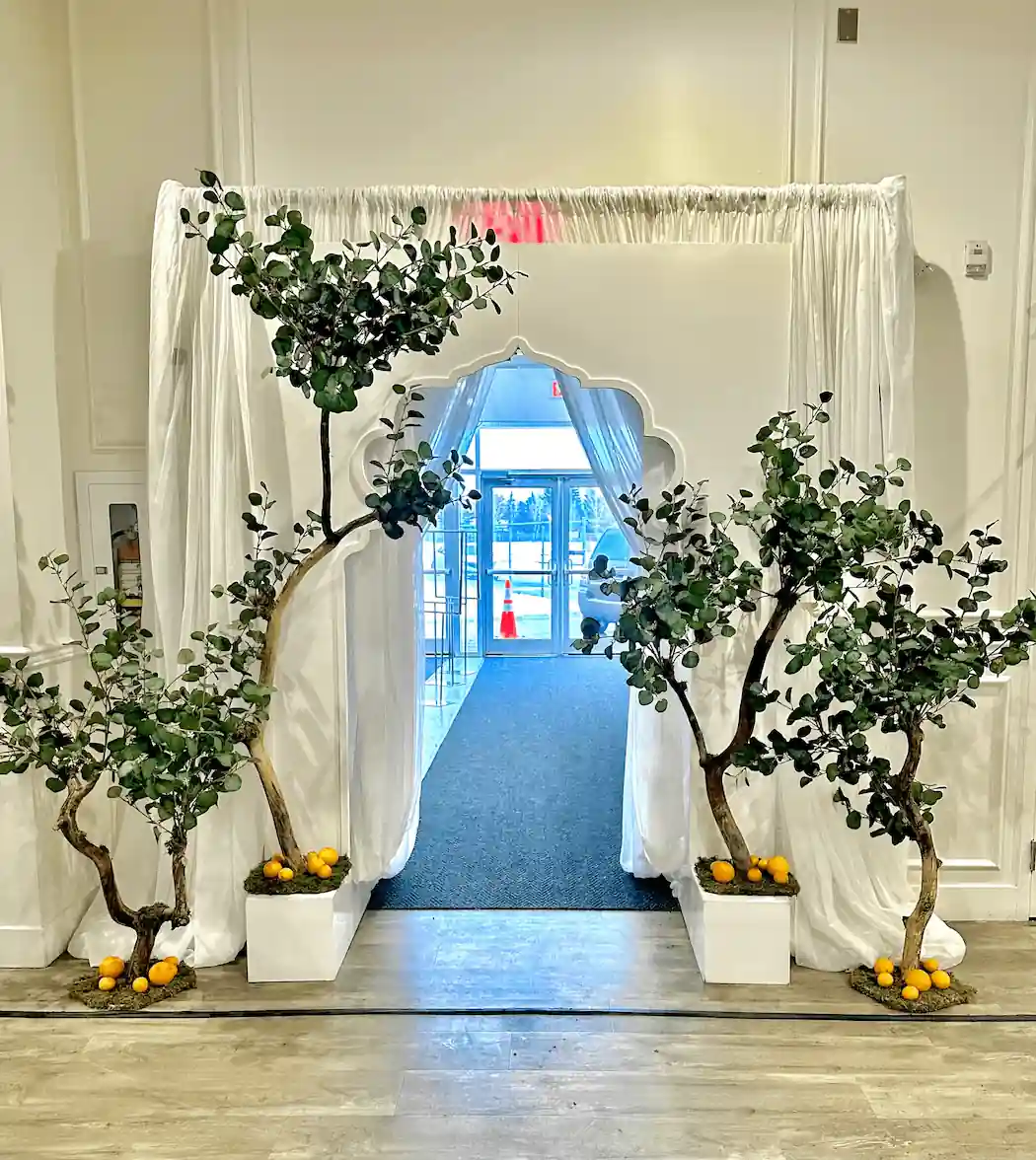 A decorative event entrance framed by white drapery and an arched cutout, flanked by potted green trees with oranges at their base. The view beyond reveals a brightly lit venue space, enhancing the welcoming atmosphere.