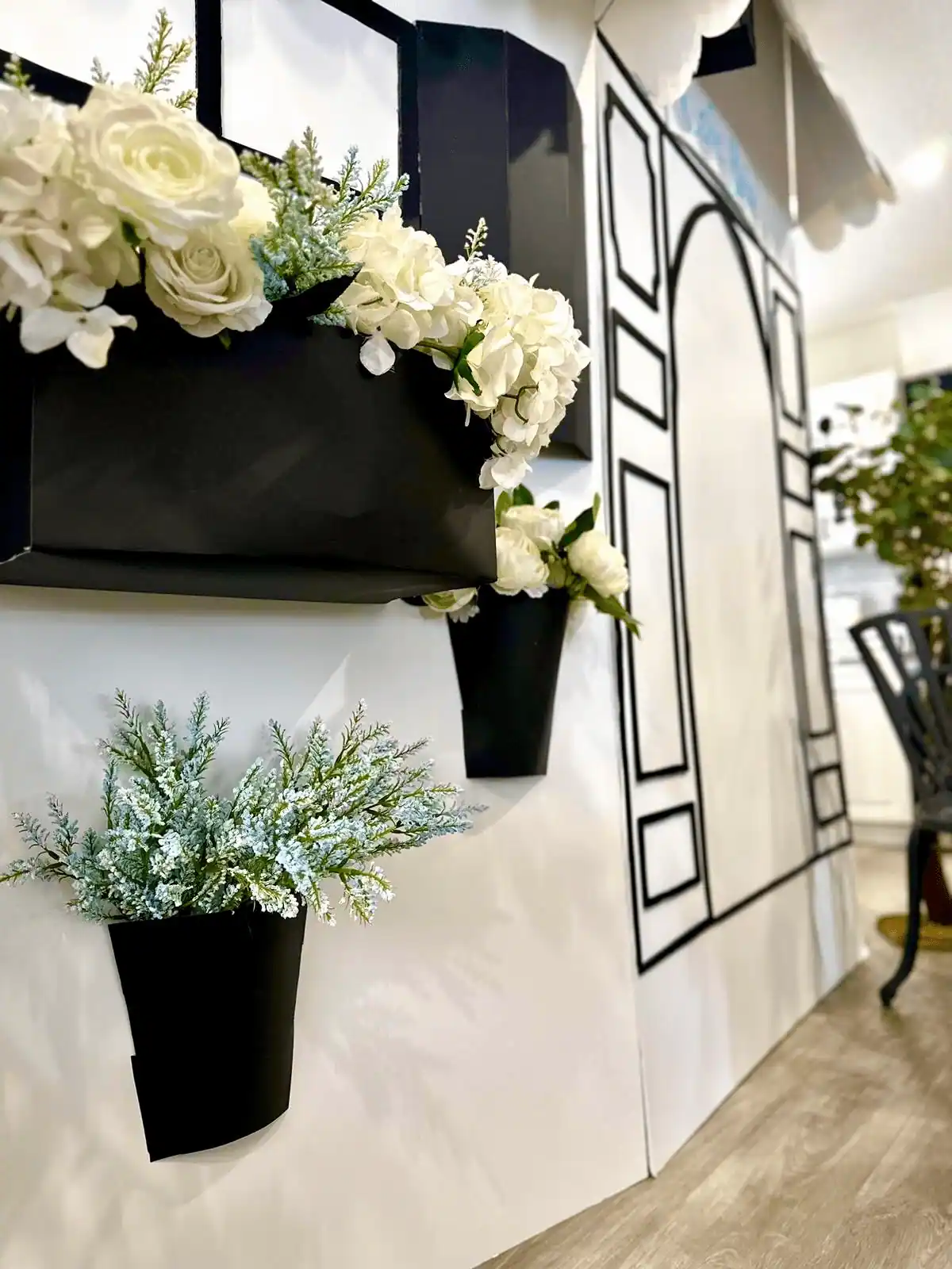 A side view of the Parisian café backdrop showcasing black window boxes filled with lush white roses and hydrangeas, accompanied by black wall-mounted planters holding frosty greenery for a sophisticated, elegant ambiance.