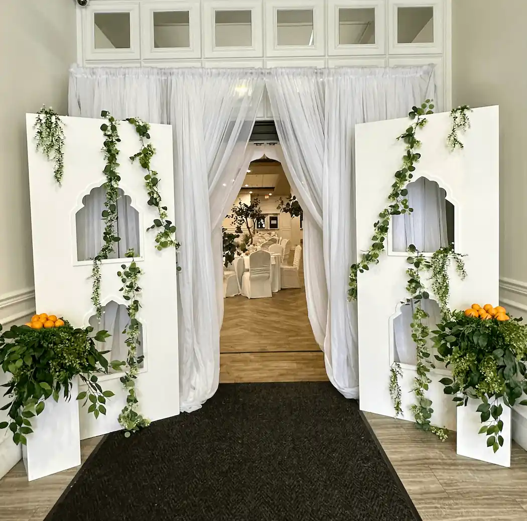 A stylish event entrance featuring tall white panels with intricate arched cutouts, adorned with cascading greenery and small orange arrangements, leading into an elegant banquet hall with white-draped chairs and lush décor.
