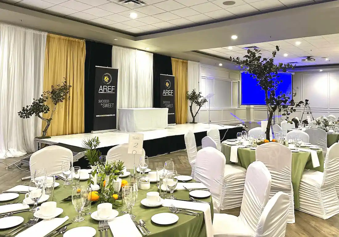 A well-decorated event venue featuring round tables draped in sage green tablecloths, set with fine china and silverware. The background showcases a modern stage with golden and white drapery, banners reading AREF SUCCESS IS SWEET, and trees adding an organic touch.