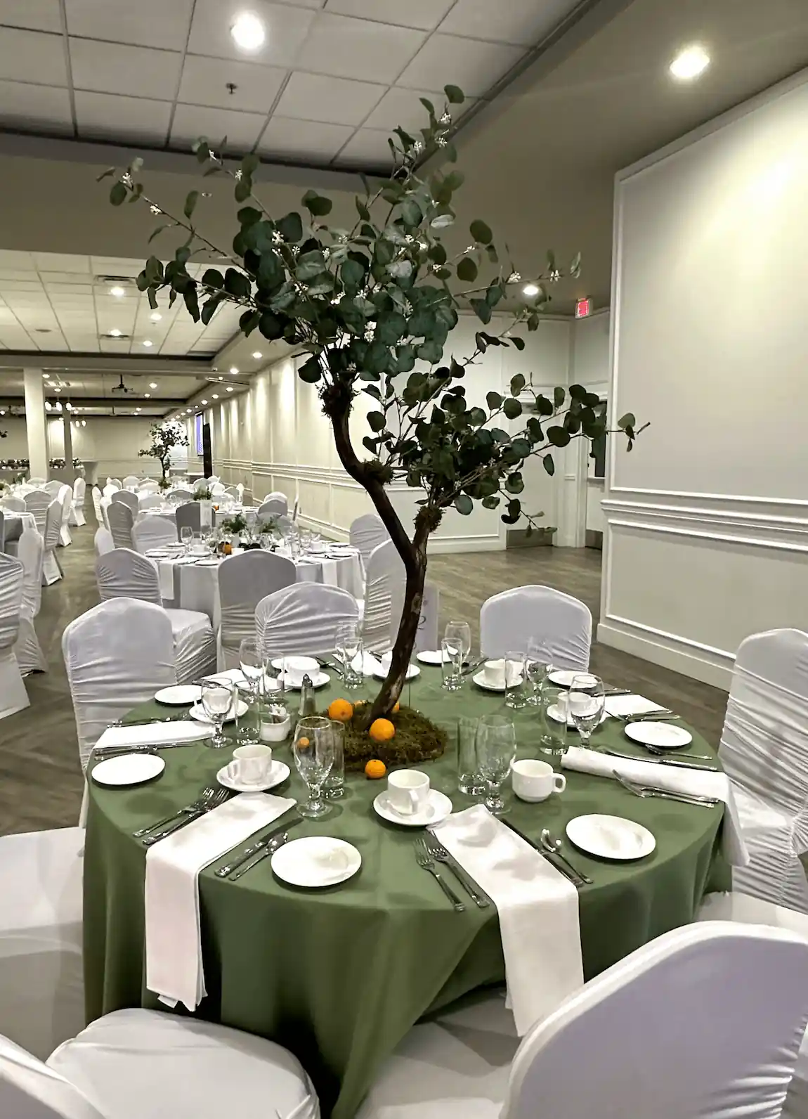 A beautifully arranged banquet table featuring a large artificial tree centerpiece set on a moss base, accompanied by scattered oranges and elegant table settings, blending nature-inspired décor with formal event styling.