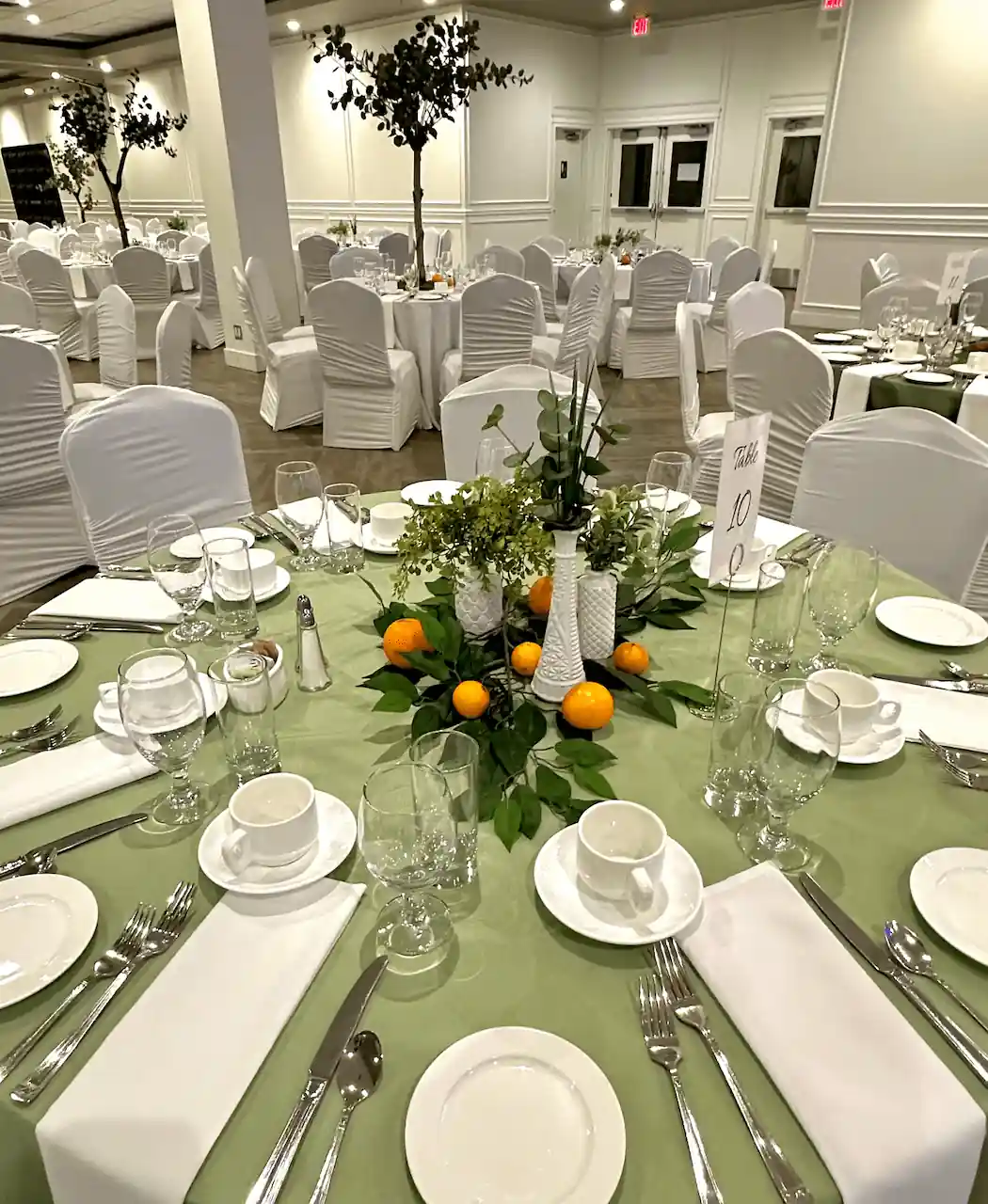 A beautifully set banquet table draped in sage green with white dinnerware, polished silverware, and an organic centerpiece featuring fresh greenery, white vases, and scattered oranges, creating a sophisticated natural aesthetic.
