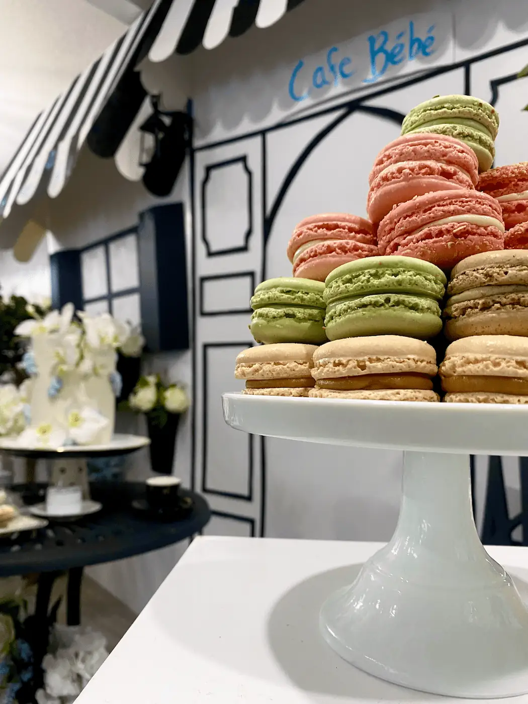 A delicate tower of pastel-colored macarons, including pistachio green, raspberry pink, and vanilla, elegantly arranged on a white cake stand with the hand-drawn Café Bébé storefront backdrop adding a charming Parisian touch.