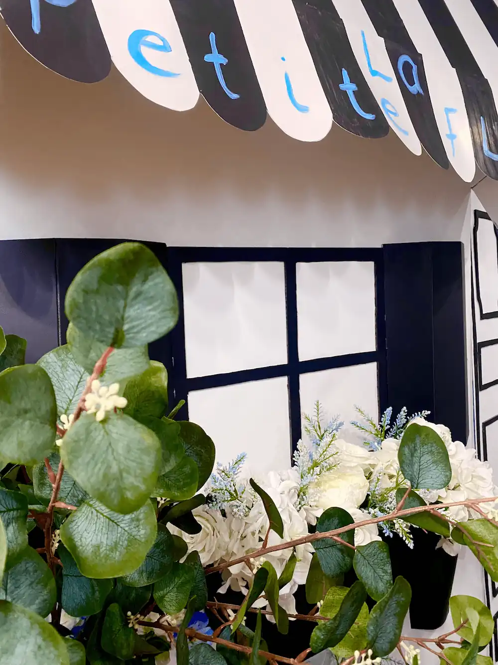 A close-up of the La Petite Fleur floral shop-inspired event setup, featuring a black-and-white striped awning, elegant window boxes filled with white hydrangeas and roses, and lush green leaves adding a natural ambiance.