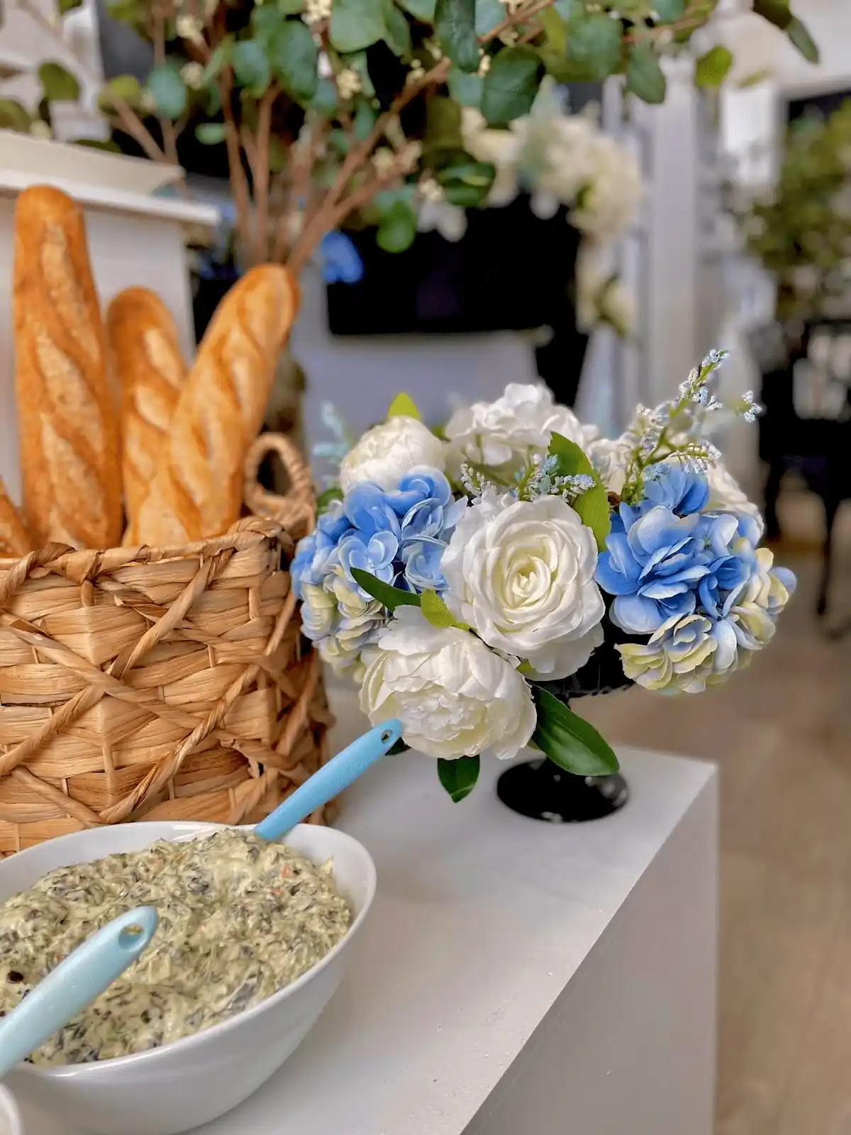 A charming bakery-style setup featuring a woven basket filled with freshly baked baguettes, a bowl of creamy dip with blue-handled serving spoons, and a floral centerpiece with white and blue roses, set against a backdrop of greenery.