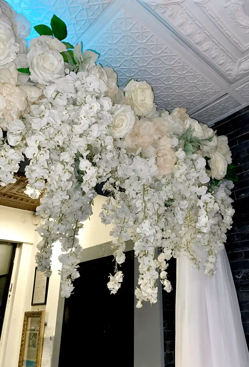A breathtaking ceiling floral arrangement with cascading white roses, wisteria, and hydrangeas, suspended from an ornate ceiling with ambient lighting, creating an elegant and romantic event atmosphere.