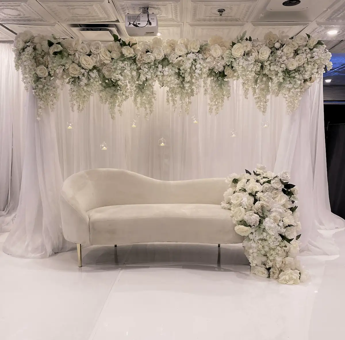 A grand event stage featuring a plush ivory chaise lounge set against a backdrop of flowing white drapery, adorned with cascading white roses, hydrangeas, and hanging glass candle orbs for a dreamy, luxurious setting.