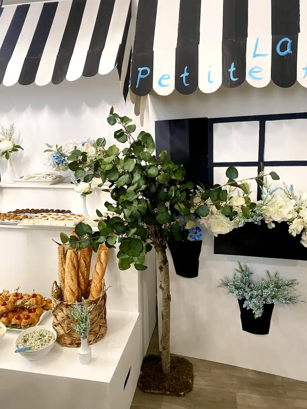 A stylish bakery-themed event setup featuring a woven basket of freshly baked baguettes, a table adorned with blue and white floral arrangements, and elegant black window boxes filled with hydrangeas.