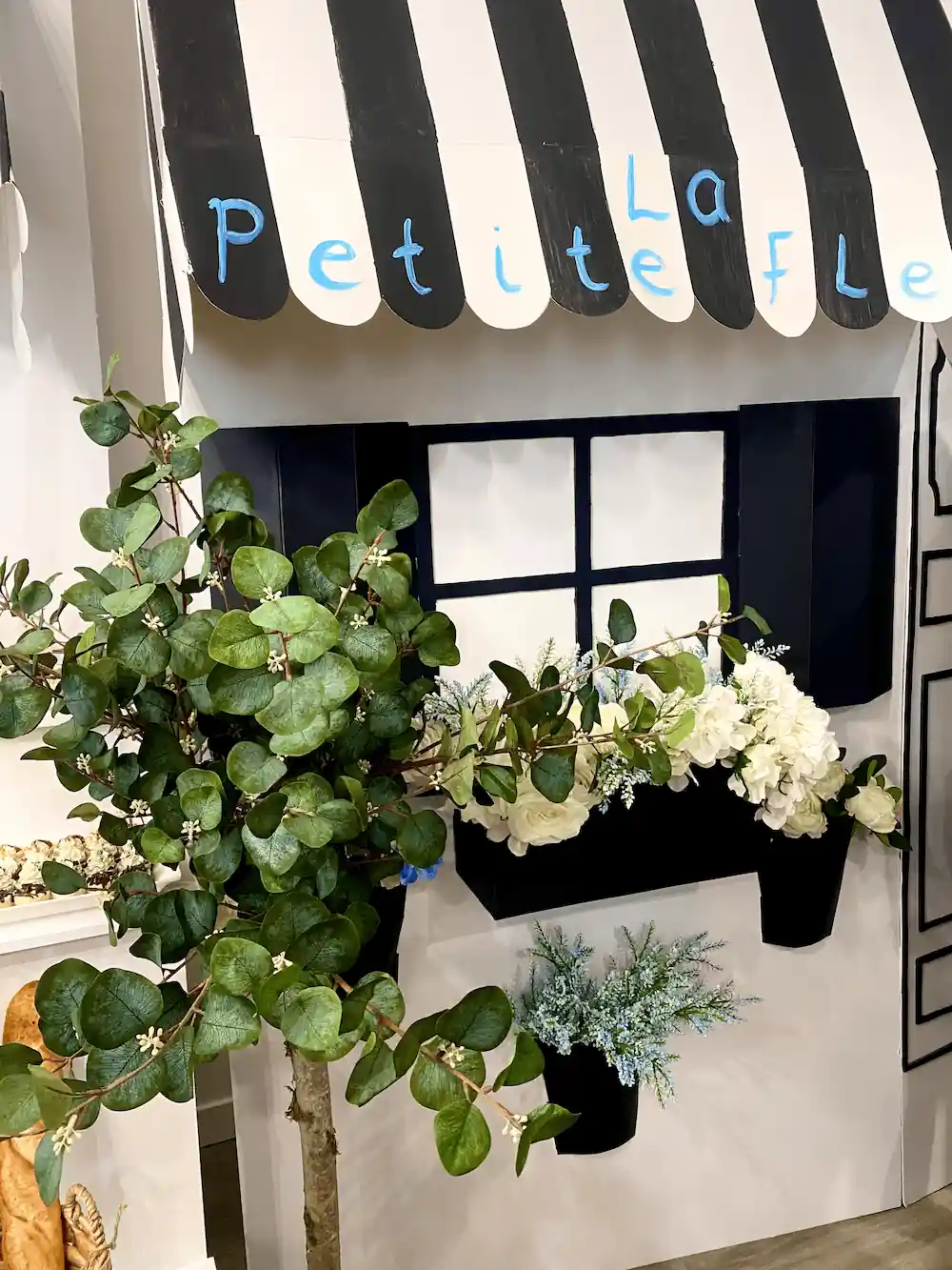 A stylish La Petite Fleur storefront setup with black-and-white striped awnings, window boxes filled with white roses and hydrangeas, and a lush artificial eucalyptus tree, creating a chic European ambiance.