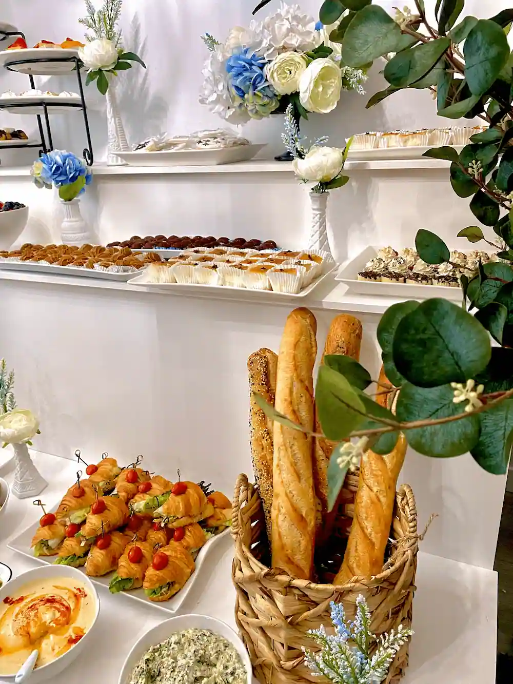 A luxurious catering display featuring freshly baked baguettes in a woven basket, croissant sandwiches with cherry tomatoes, creamy dips, and an assortment of pastries, beautifully arranged with white and blue floral accents.