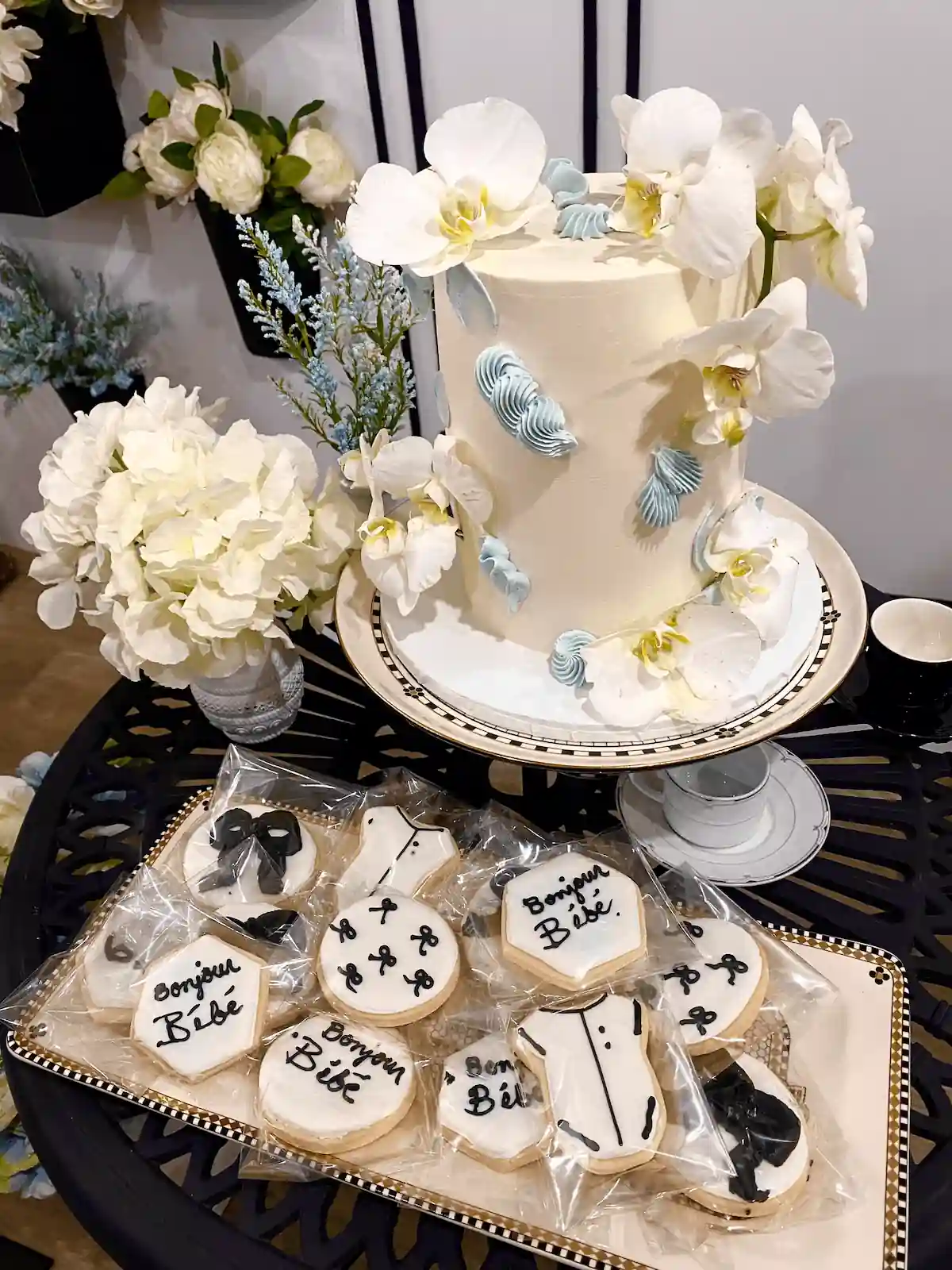 A two-tier vanilla cake adorned with delicate white orchids and light blue buttercream accents, paired with intricately designed cookies reading Bonjour Bébé, all displayed on a vintage-inspired black lace table.