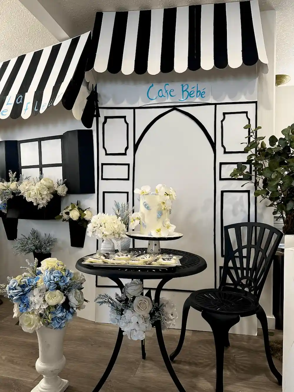 A black-and-white Café Bébé storefront backdrop featuring a vintage-style table set with a floral-adorned cake, themed sugar cookies, and elegant white and blue floral accents.