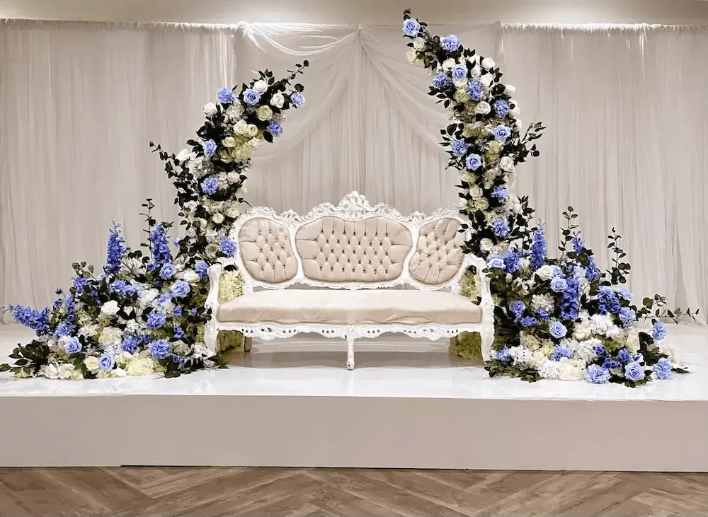 Luxurious bridal shower stage setup featuring an ornate vintage ivory chaise lounge with tufted upholstery, framed by elegant cascading floral arrangements of white and soft blue roses, hydrangeas, and lush greenery. The backdrop is draped in sheer white fabric, creating a sophisticated and romantic ambiance perfect for weddings and special events