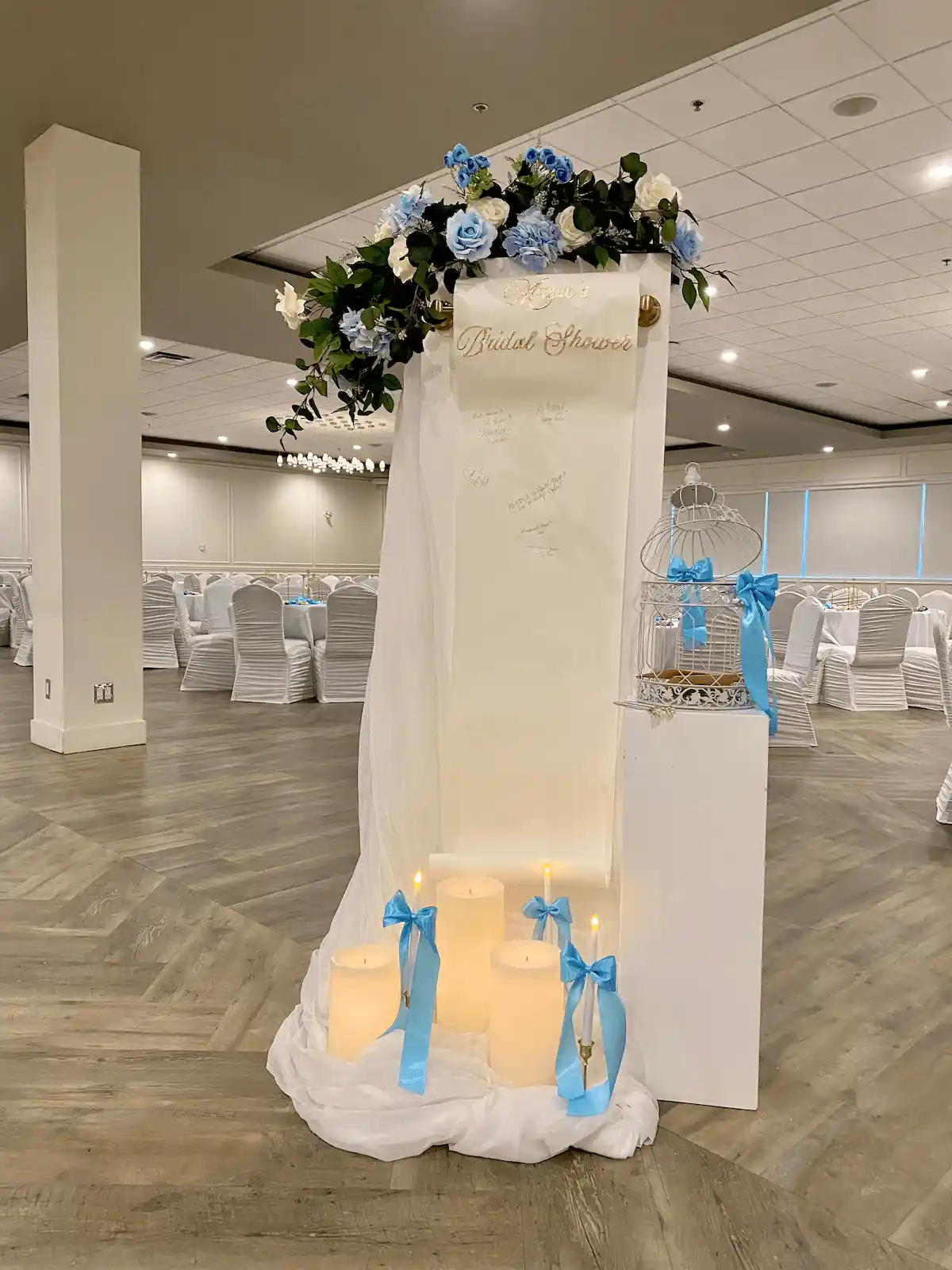 Bridal shower guestbook station with a custom scroll-style sign, floral accents, and glowing LED candles wrapped in soft blue ribbons. Elegant and romantic wedding decor idea for guest messages and well wishes.