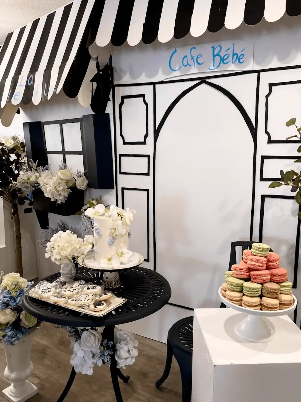 A beautifully styled Parisian-themed dessert table with a black-and-white striped awning labeled Café Bébé, featuring a white floral cake, a plate of decorated cookies, and a tall white pedestal displaying colorful macarons and a croquembouche.