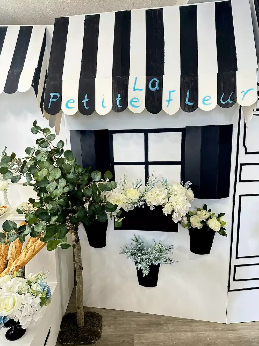A close-up of the La Petite Fleur flower shop-inspired backdrop with a black-and-white striped awning, black window boxes overflowing with white and green floral arrangements, and a small artificial tree adding to the Parisian ambiance.