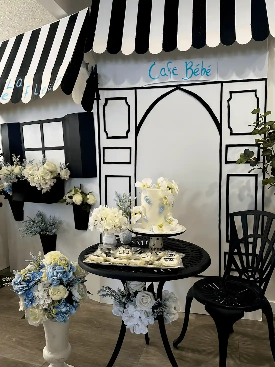 A full view of the Café Bébé and La Petite Fleur event setup, featuring a black bistro table with an elegant cake display, a charming Parisian-style storefront backdrop, and a buffet table filled with pastries and desserts, creating a cozy French café atmosphere.