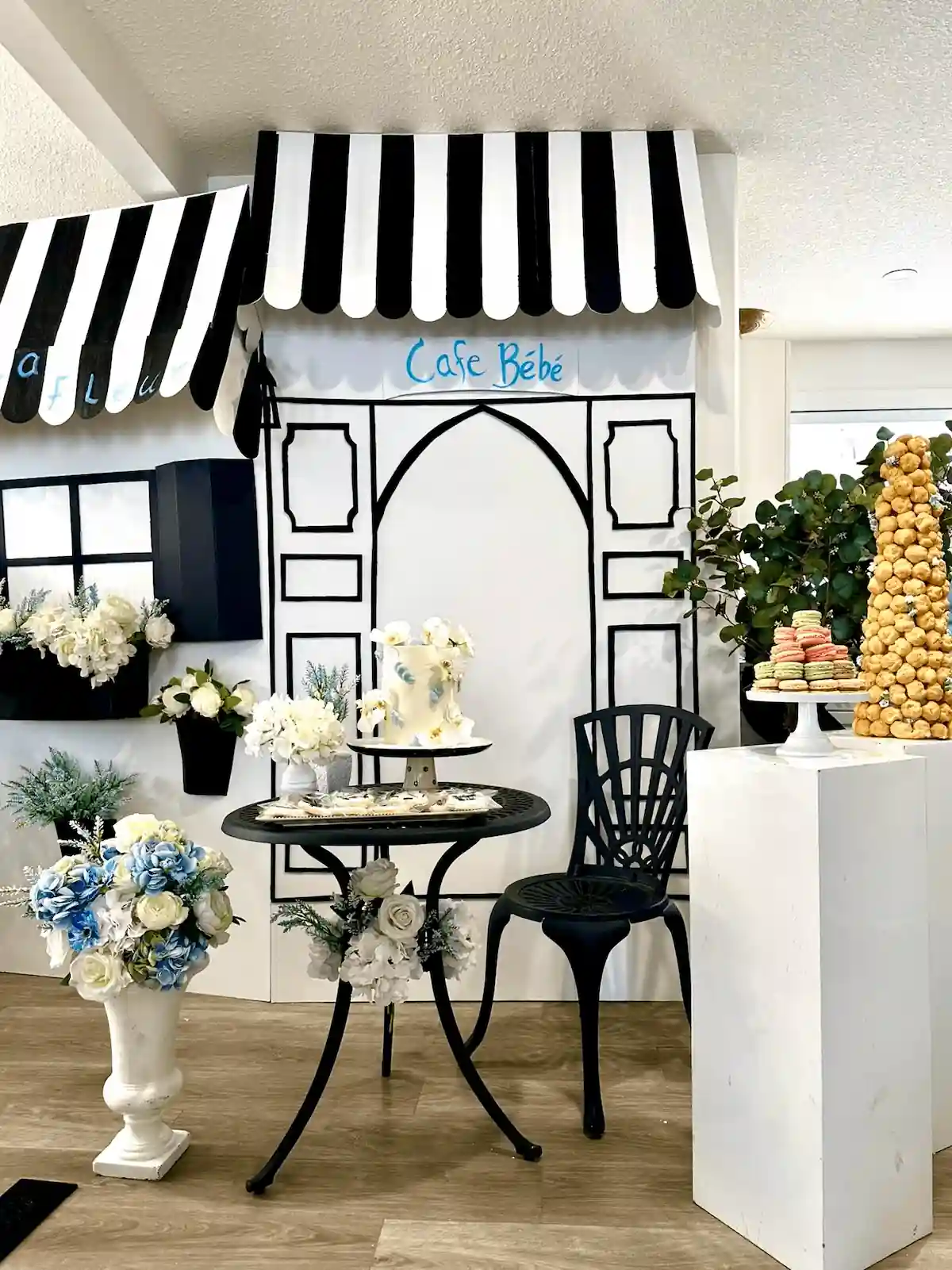 A charming café-themed event setup features a black-and-white striped awning with Café Bébé written above an illustrated storefront backdrop, complemented by a small black bistro table with floral decorations, a matching chair, and an elegant cake display surrounded by white and blue floral arrangements, macarons, and a towering croquembouche.