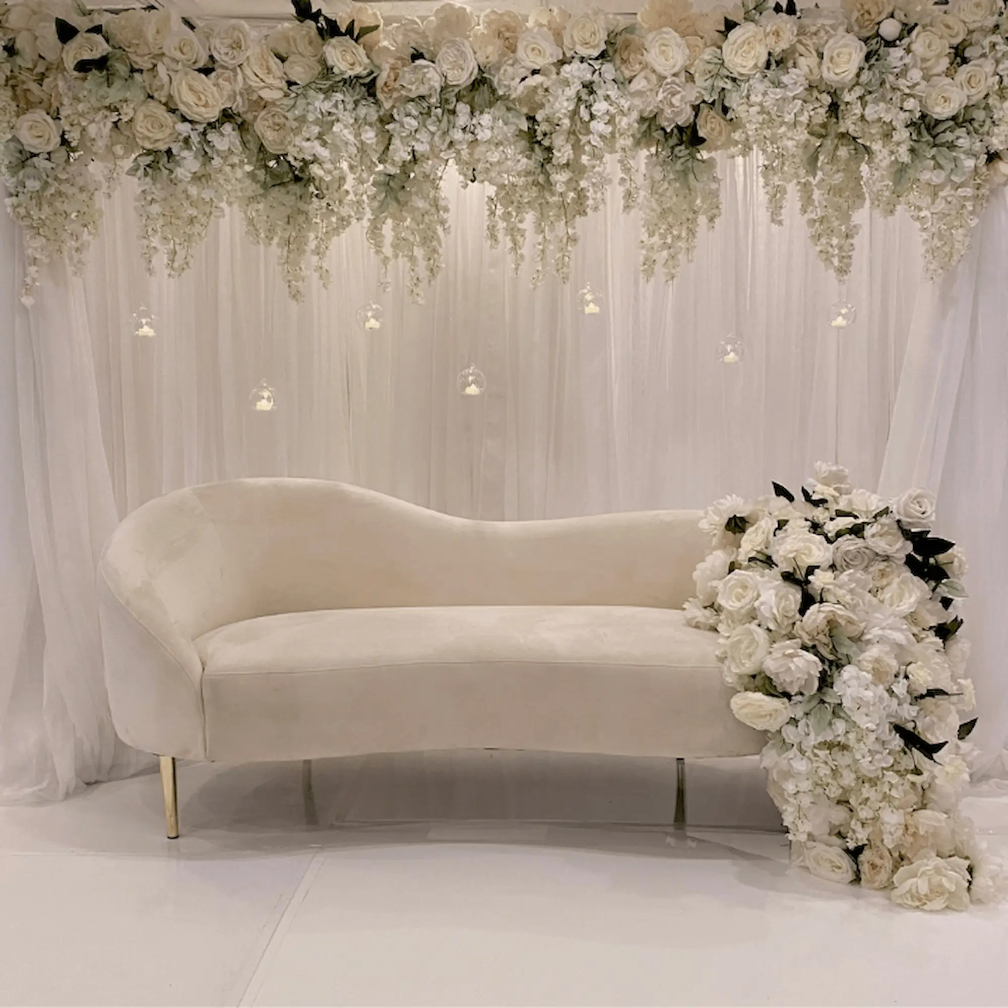 Elegant event decor featuring a plush ivory chaise lounge surrounded by cascading white floral arrangements and delicate hanging votive candles. The scene is set against a backdrop of sheer white drapery, exuding sophistication and luxury.
