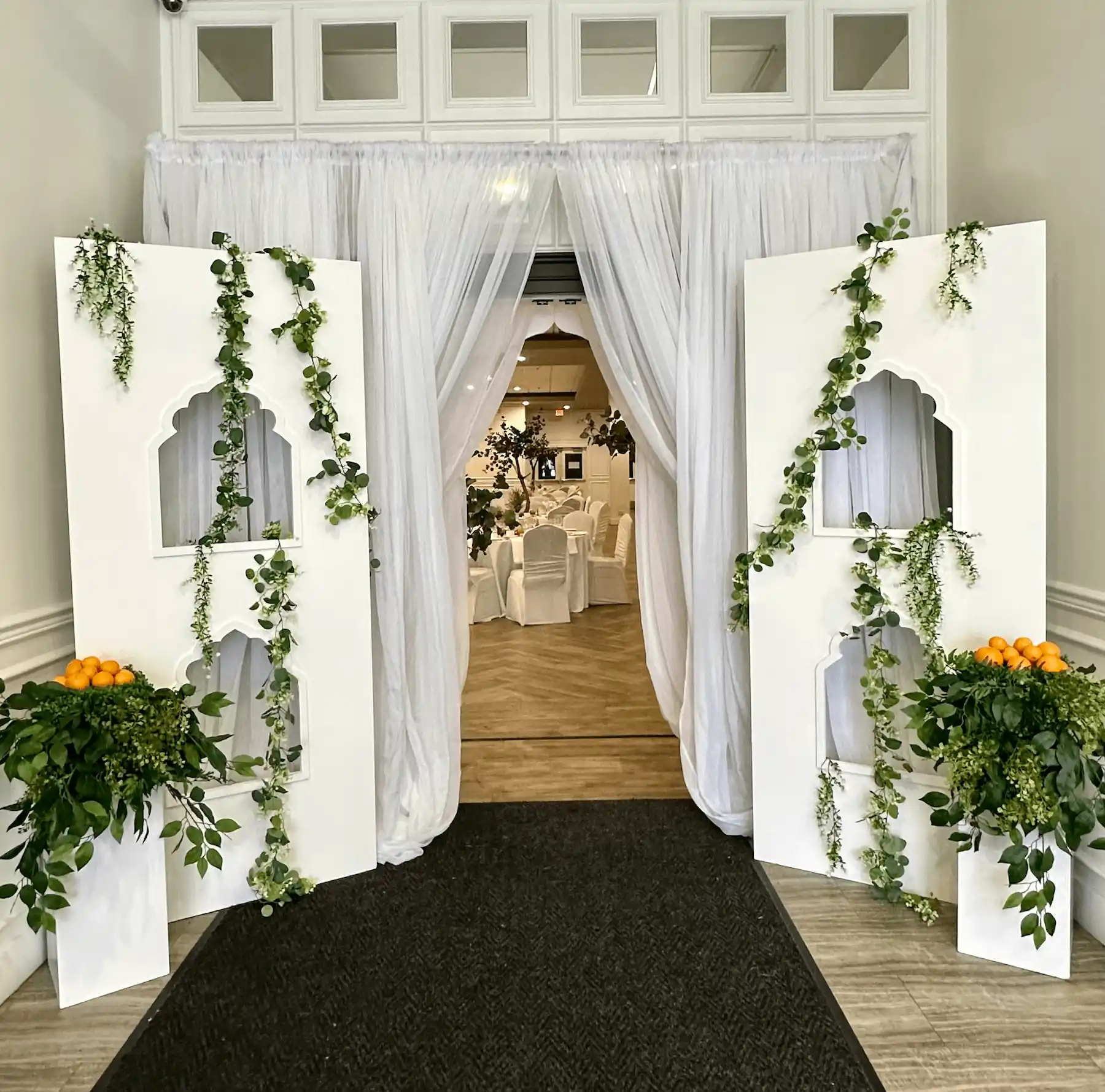 An elegant event entrance with white drapery and decorative white panels featuring arched cutouts, adorned with cascading green vines. The panels are flanked by lush greenery and small bunches of oranges. Through the opening, a banquet hall with round tables covered in white linens and chairs is visible, decorated with greenery and warm lighting.