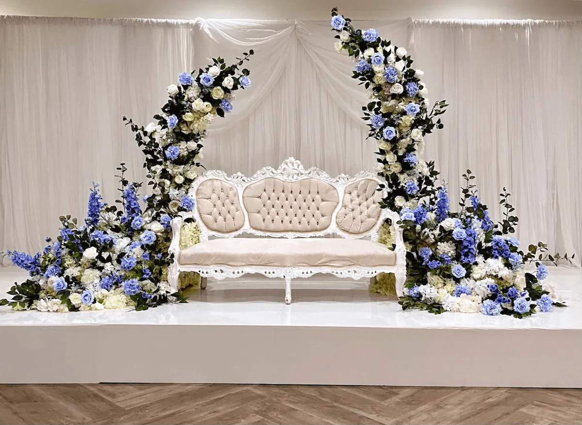 Elegant bridal shower stage with a vintage tufted sofa, surrounded by cascading floral arches featuring white, ivory, and soft blue roses, hydrangeas, and greenery. Luxurious wedding backdrop with draped white fabric for a sophisticated event decor.