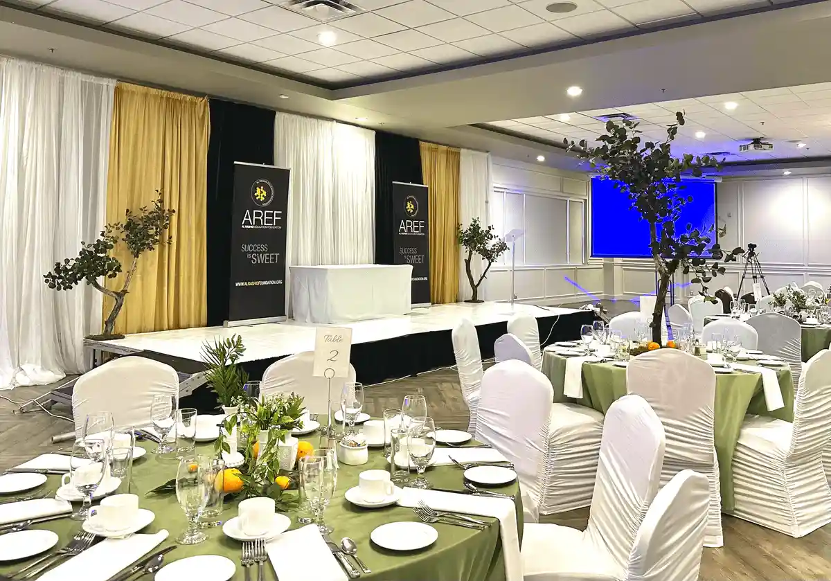 Banquet hall with neatly arranged tables featuring green tablecloths, citrus-themed centerpieces, and a staged area with banners and lighting for an event.