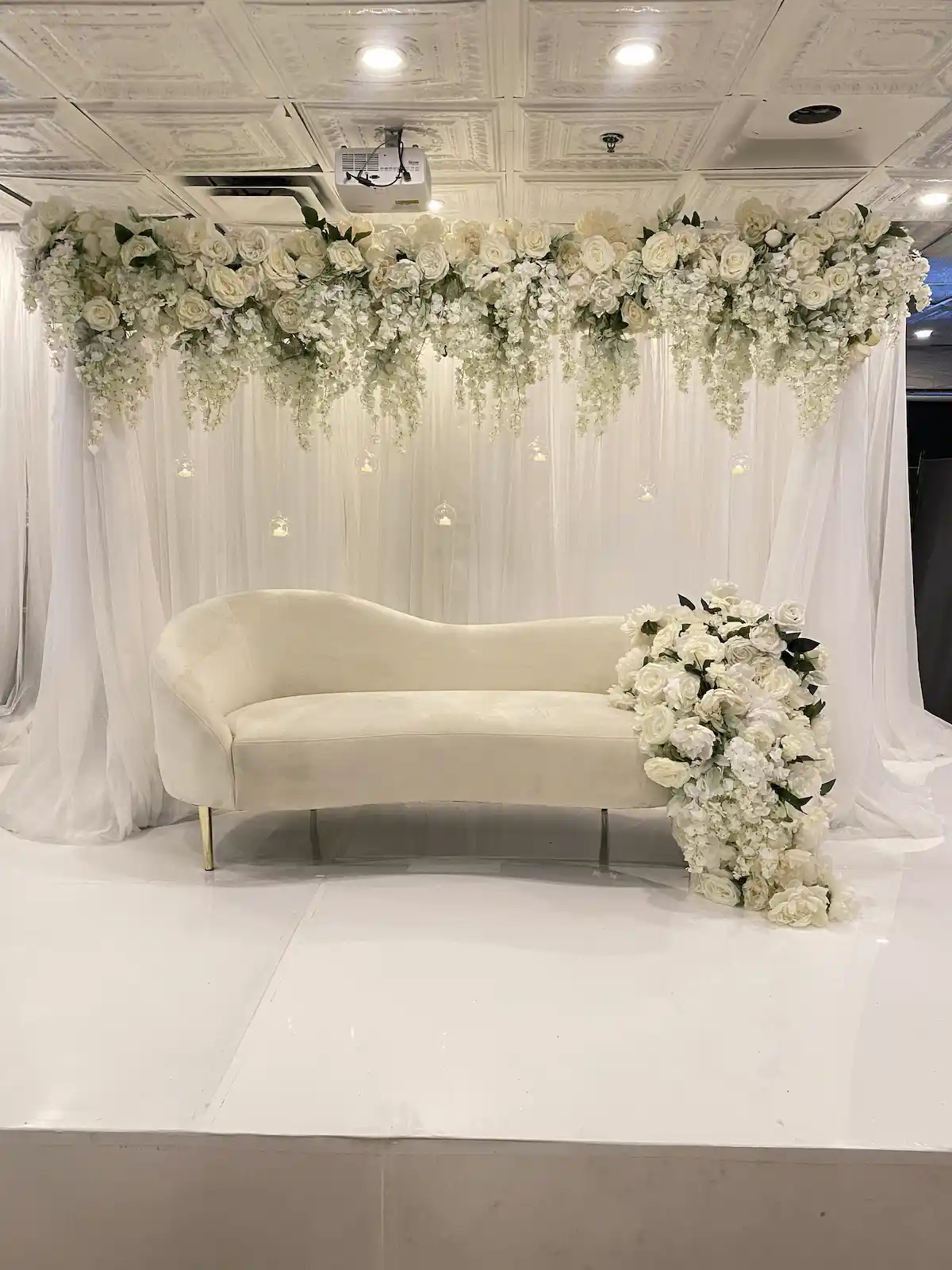 A luxurious event stage features a plush ivory chaise lounge set against a backdrop of flowing white drapery, with cascading white roses and hydrangeas adorning the top and side, complemented by delicate hanging glass orbs, creating an elegant and romantic ambiance.